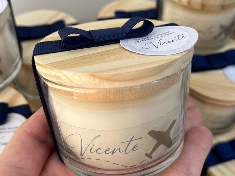 Vela em copo de vidro transparente com a cera creme vis�vel e tampa de madeira clara encaixada, la�o de gorgur�o azul escuro passando por baixo da tampa