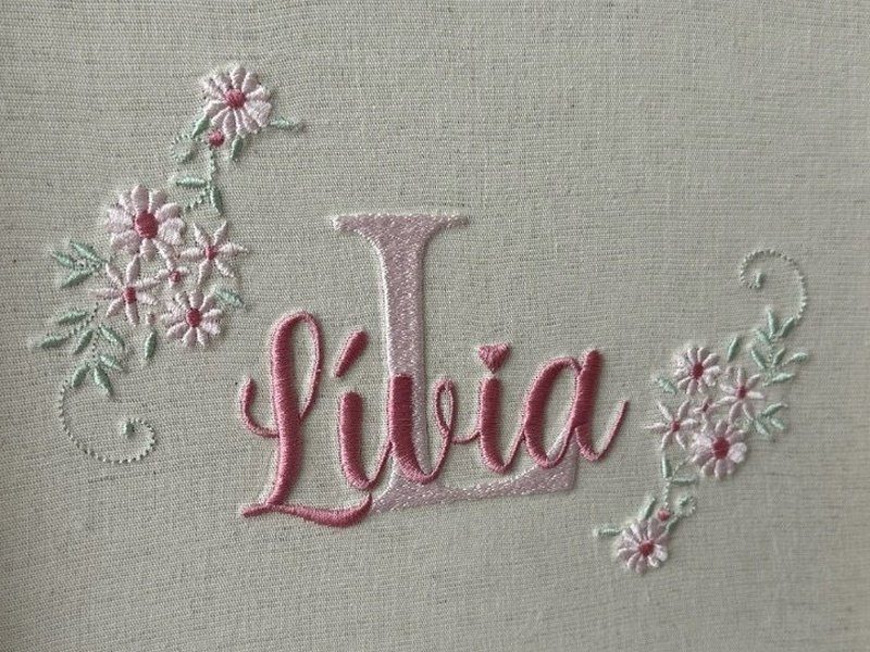 O monograma inteiro: a letra L grande em rosa claro atr�s do nome Livia em cursiva rosa escuro, ladeado por dois ramos de margaridas brancas de miolo rosa