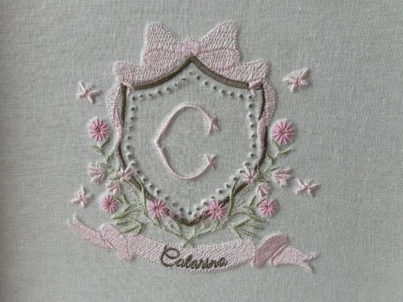 O bras�o bordado por inteiro: escudo contornado de perolinhas com a letra C, la�o rosa no topo, borboletas, margaridas e a faixa com o nome Catarina em cursiva marrom