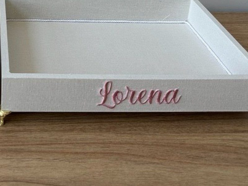 O nome Lorena bordado em cursiva rosa na lateral externa da bandeja de linho, acima dos p�s dourados