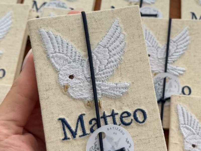 A pomba branca bordada em relevo cheio sobre o linho cru, com as asas abertas, e o nome Matteo bordado em azul escuro embaixo