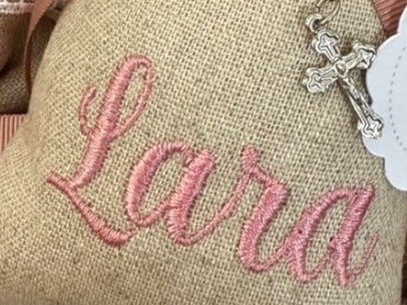 O nome Lara bordado em rosa, em cursiva grande, sobre o linho do saquinho
