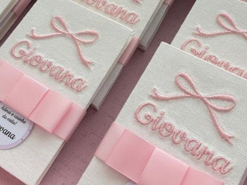 O nome Giovana bordado em cursiva rosa com relevo alto, sob um laco bordado, na capa de linho do livrinho