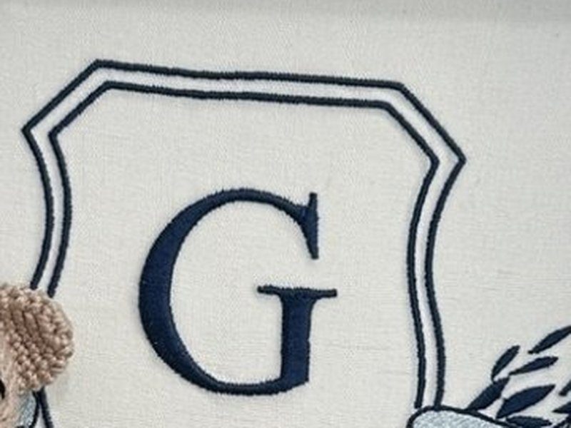 Escudo bordado em azul escuro com a letra G ao centro, contorno duplo do escudo e ramos de folhas saindo pela direita