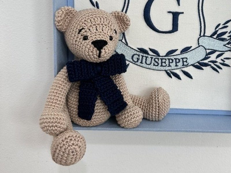 Urso de croch� bege com la�o de croch� azul escuro no pesco�o, sentado na borda inferior da caixa com as pernas para fora do quadro