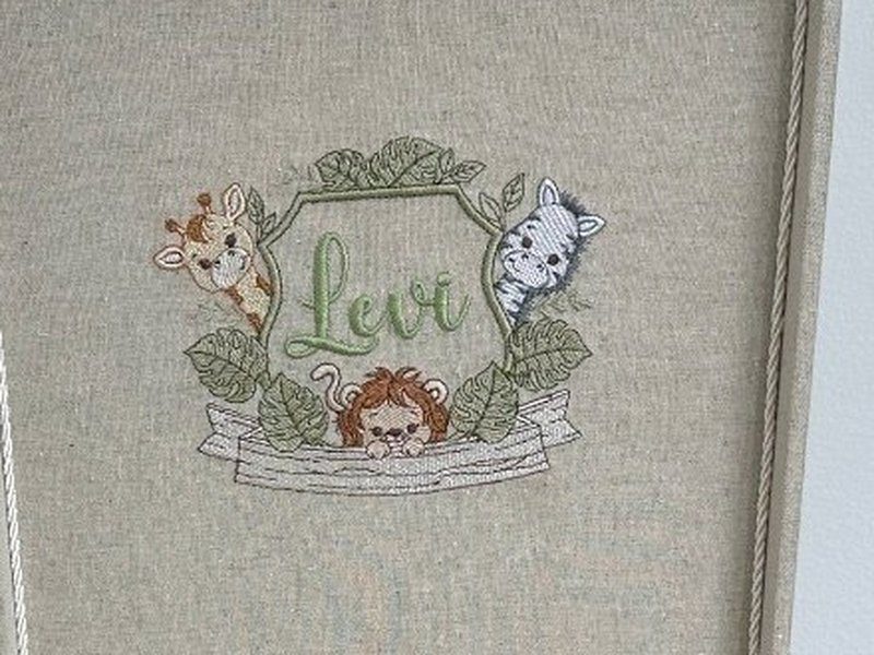 Bras�o bordado dentro da tampa: girafa e zebra espiando entre folhagens, le�o sobre a faixa e o nome Levi em verde dentro da moldura