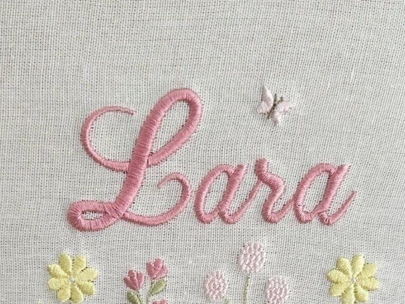 O nome Lara inteiro em cursiva rosa, com o relevo do ponto vis�vel e a trama do linho ao redor