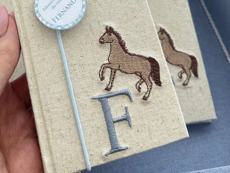 Cavalo bordado em marrom sobre a capa de linho, com a inicial F em azul logo abaixo