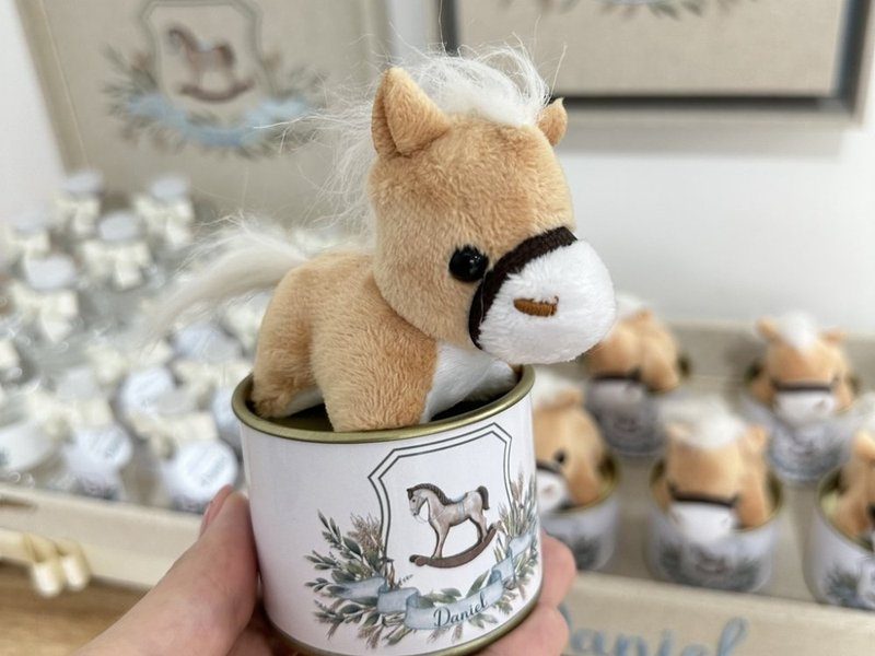Lata com potro de pel�cia bege de crina branca encaixado na boca, e r�tulo com o bras�o de cavalo de balan�o entre folhagens e o nome Daniel