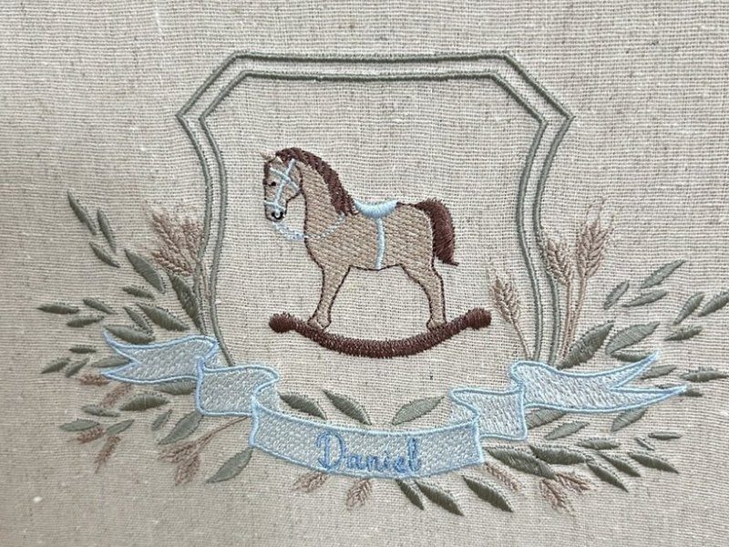 Bras�o bordado por inteiro: escudo em contorno verde, cavalo de balan�o em marrom e bege, ramos e espigas, e faixa azul-clara com Daniel