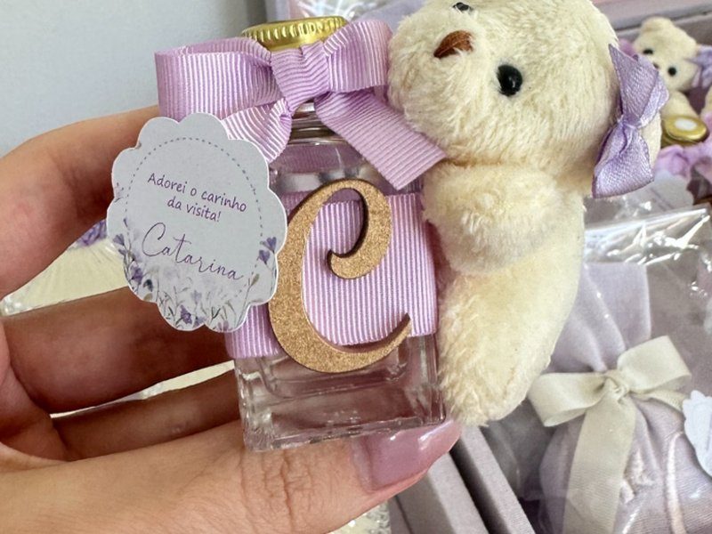 O difusor de vidro com tampa dourada, fita lil�s, a inicial C dourada aplicada, o ursinho de pel�cia com la�o lil�s e a tag rendada com 'Adorei o carinho da visita!' e o nome Catarina