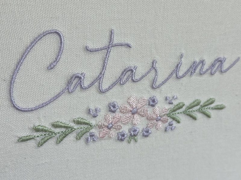 O nome Catarina bordado em cursiva lil�s sobre linho cru, com o ramo floral logo abaixo: flores lil�s e rosa claro ao centro e folhas verdes em espiga para os dois lados
