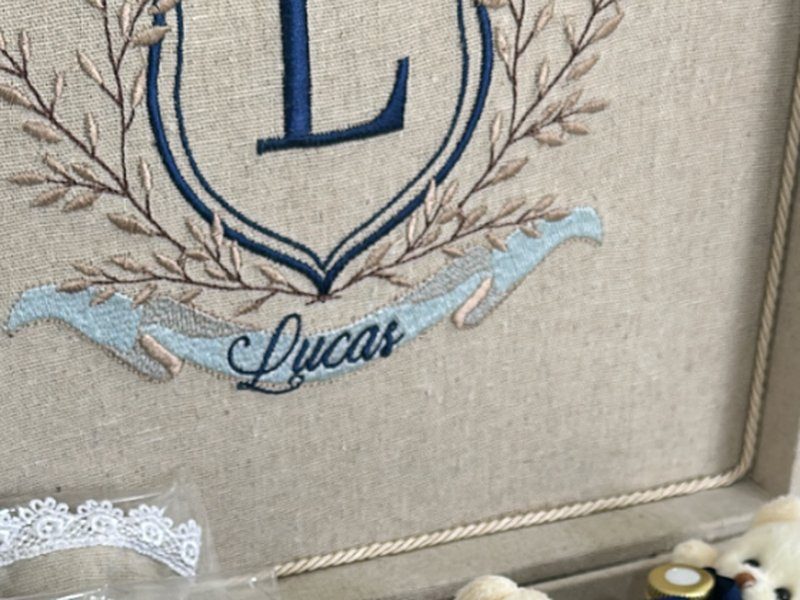Escudo com L, ramos laterais e faixa azul clara com Lucas em cursiva, bordados na tampa de linho bege