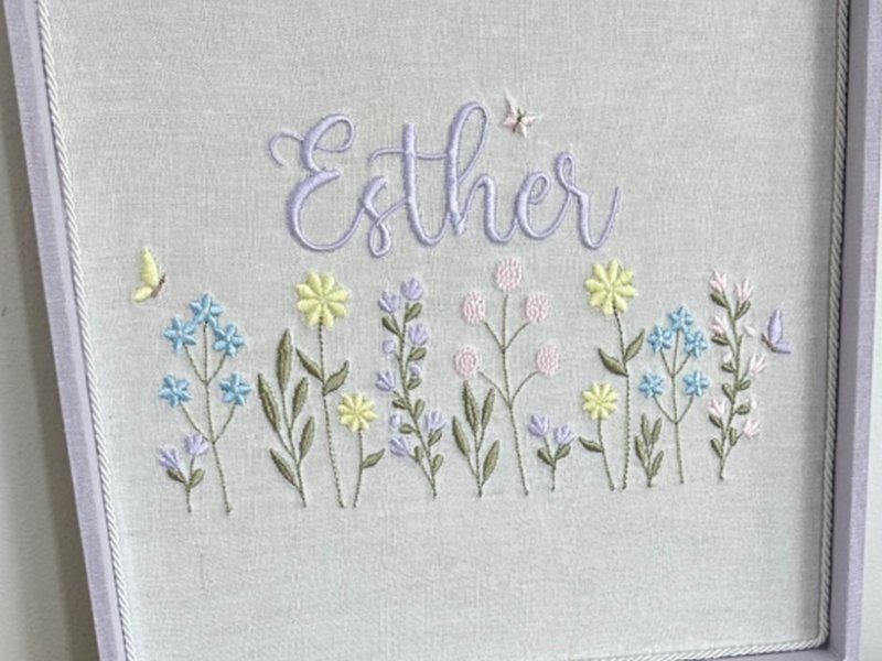 O nome Esther em cursiva lil�s sobre um jardim bordado em tons pastel, com uma borboleta amarela � esquerda