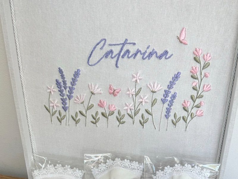 O nome Catarina em cursiva lil�s com uma borboleta rosa ao lado, sobre um jardim bordado de lavandas e flores rosa