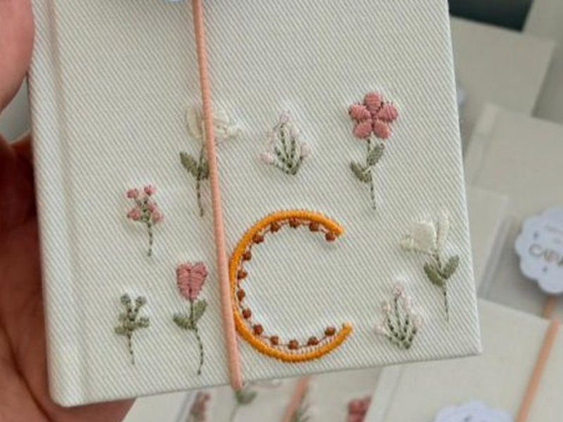 Inicial C em laranja pontilhada cercada de flores silvestres soltas bordadas em tecido de trama diagonal
