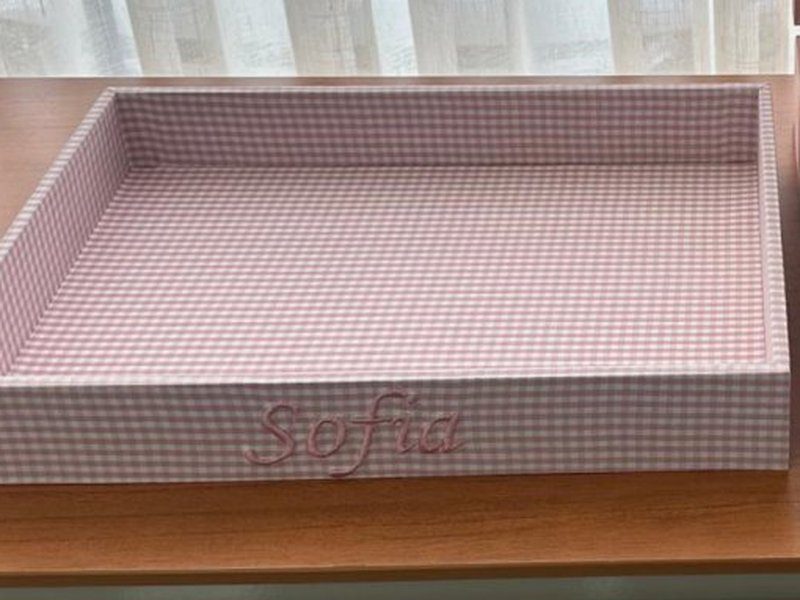 A bandeja xadrez rosa com o nome bordado na frente, ao lado da caixa de linho rosa aberta
