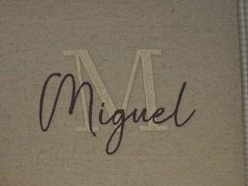 Inicial M grande em bege com o nome Miguel em cursiva escura sobreposto