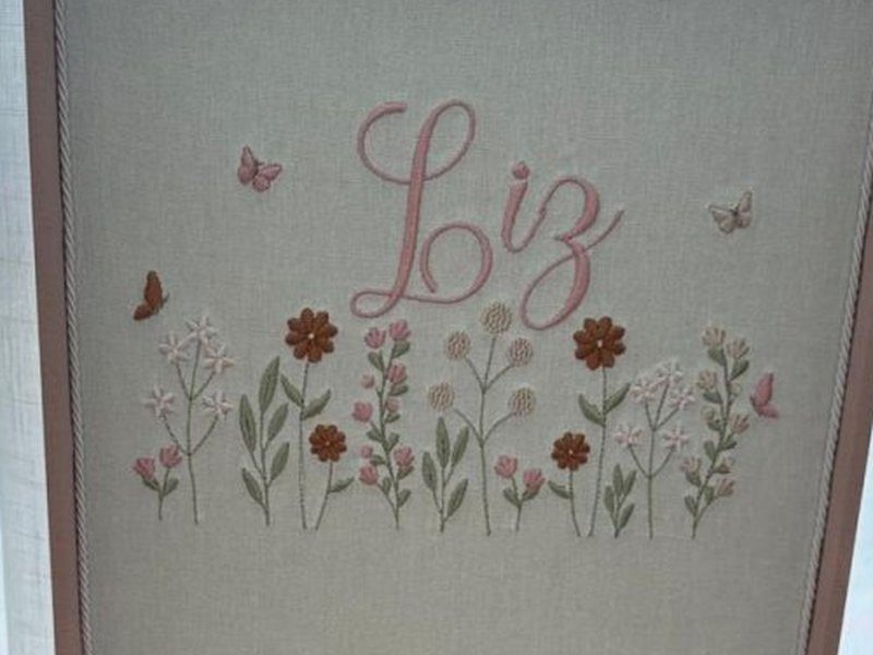 Fileira de flores bordadas (margaridas, trevos e bot�es) com Liz em cursiva rosa acima e borboletas