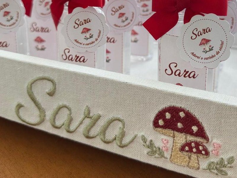 Sara em cursiva verde-sage ao lado de dois cogumelos bordados de chap�u vermelho com pintas brancas