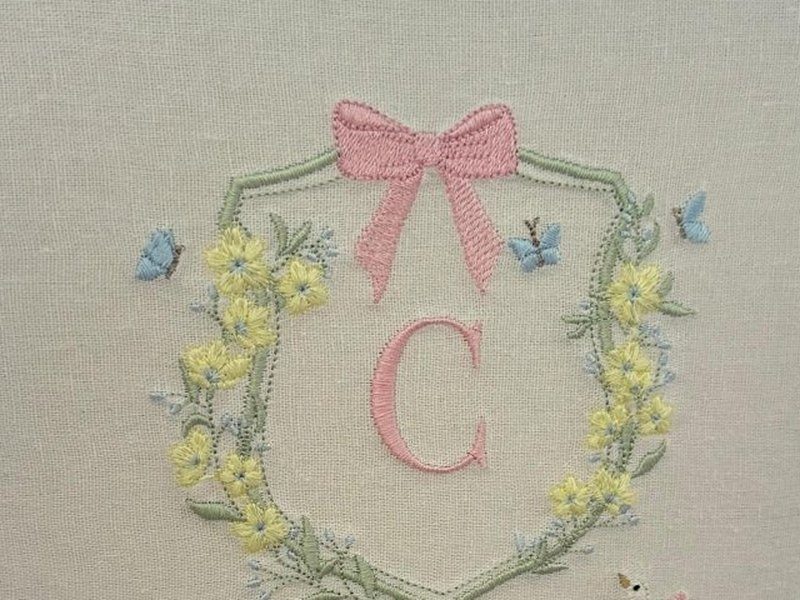 O escudo bordado com la�o rosa no topo, a inicial C rosa ao centro e a coroa de flores amarelas com folhagem verde