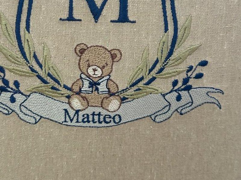 O ursinho marrom de gola de marinheiro sentado sobre a faixa clara onde o nome Matteo est� bordado