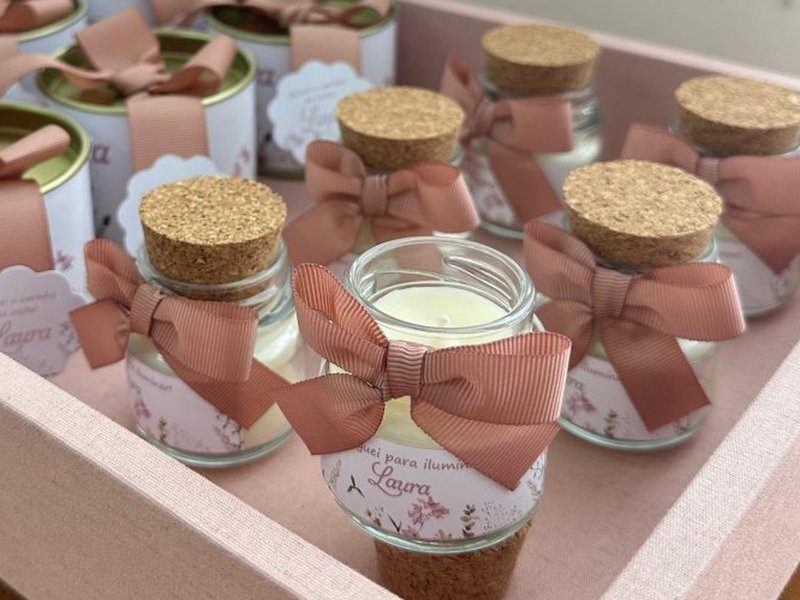 Potes de vidro com rolha de corti�a e la�o rose, um deles aberto mostrando a vela; latas douradas com la�o ao lado