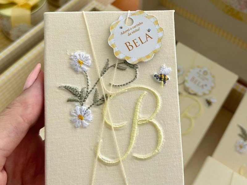 Caixinha-livro com o monograma B bordado em amarelo, margaridas brancas de miolo laranja em relevo e uma abelha aplicada