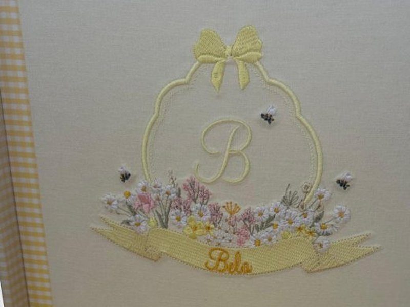 Moldura ondulada bordada com la�o amarelo, inicial B, campo de flores silvestres coloridas, abelhas e faixa amarela com o nome Bela