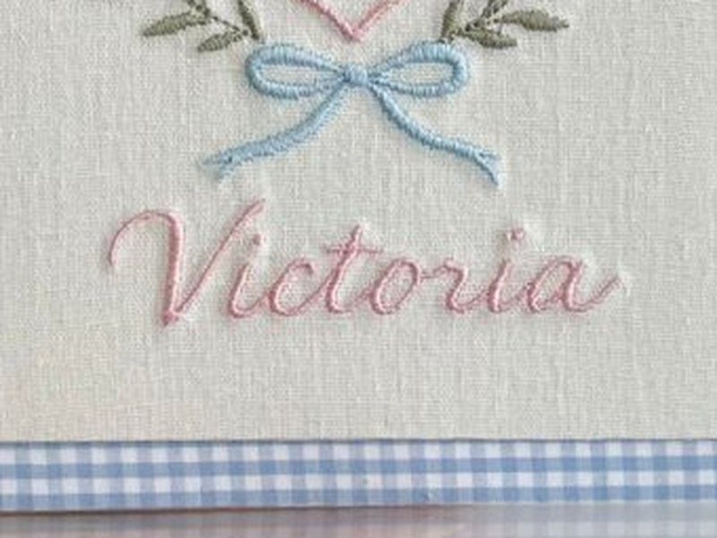 O nome Victoria bordado por inteiro em cursiva rosa, abaixo do escudo