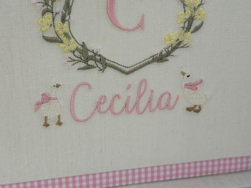 Dois gansos brancos de gorro ladeando o nome Cec�lia bordado em cursiva rosa