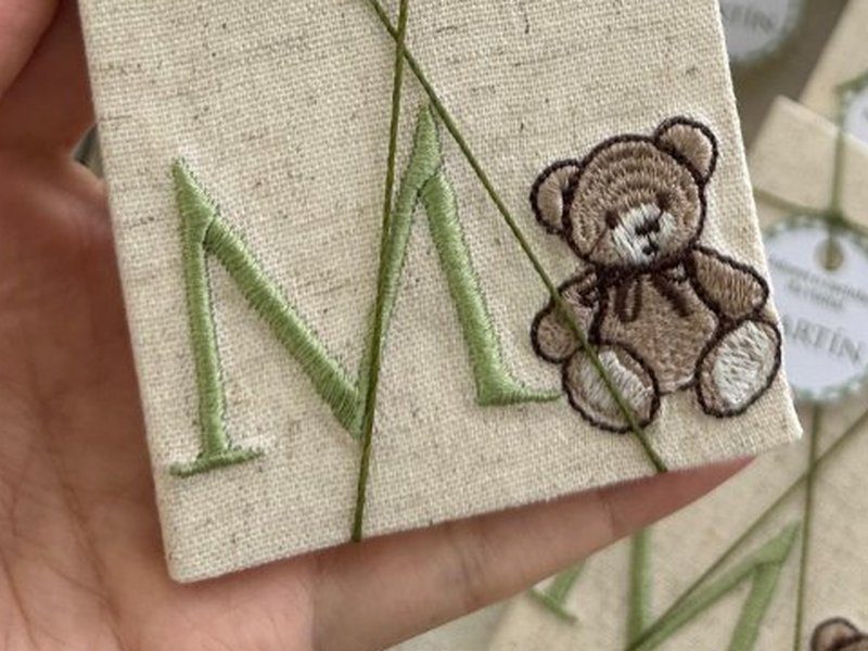 Caixinha-livro com a inicial M bordada em verde e um ursinho bordado em marrom apoiado nela