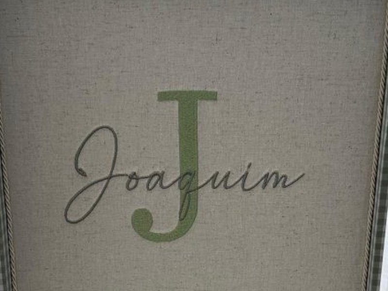 Letra J verde grande com Joaquim em cursiva cinza escrita por cima, bordados no linho da parte interna da tampa