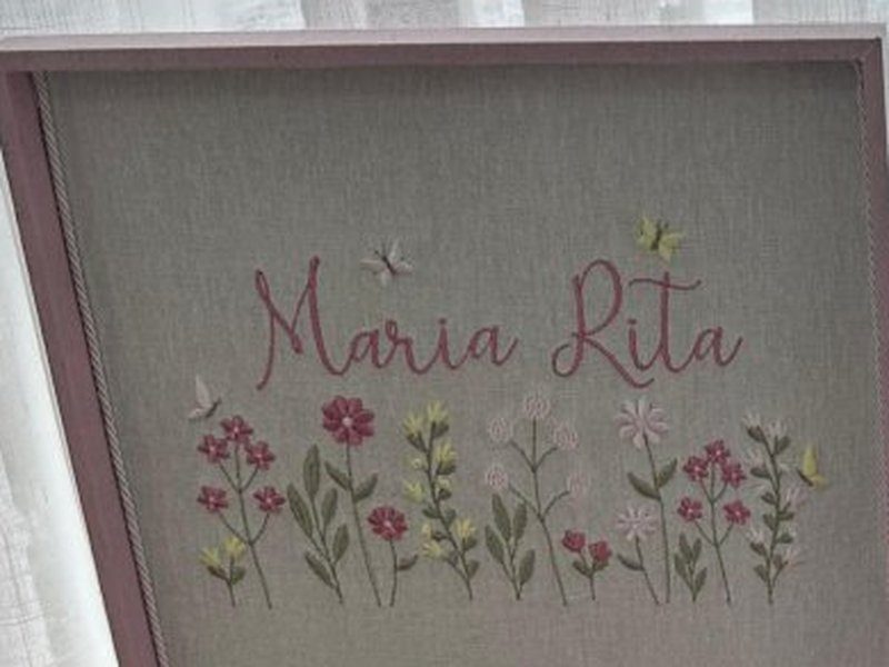 'Maria Rita' em cursiva rosa e o jardim bordado logo abaixo, com flores rosas, amarelas e brancas e borboletas, sobre linho cinza-taupe