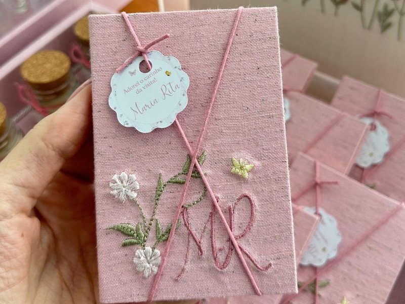 Capa de linho rosa com 'MR' em cursiva, margaridas brancas e folhas verdes, cord�o rosa cruzado e tag rendada