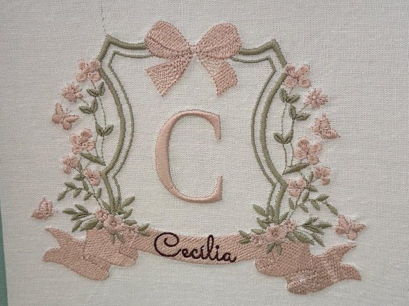 O bras�o inteiro: escudo verde escuro, la�o rosa no topo, coroa de flores e borboletas, letra C e faixa com 'Cec�lia' em cursiva escura