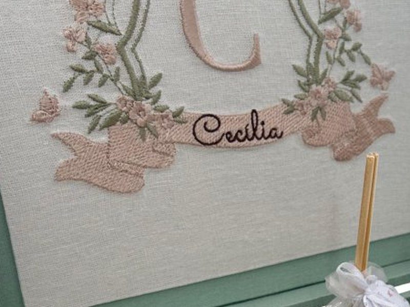 'Cec�lia' em cursiva escura bordado sobre a faixa rosa, entre as pontas enroladas da fita
