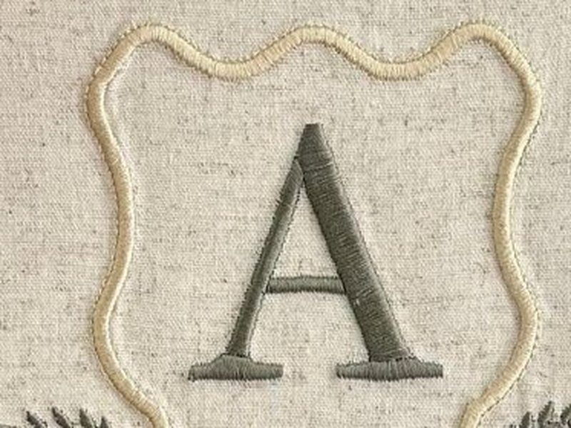 Escudo de contorno creme ondulado, feito em cord�o, com a letra A verde escuro em ponto cheio ao centro