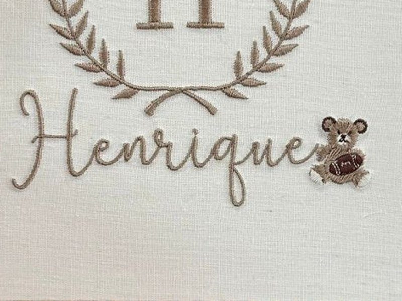 O nome Henrique bordado por inteiro em cursiva bege, com um ursinho marrom pequeno sentado � direita, encostado na �ltima letra
