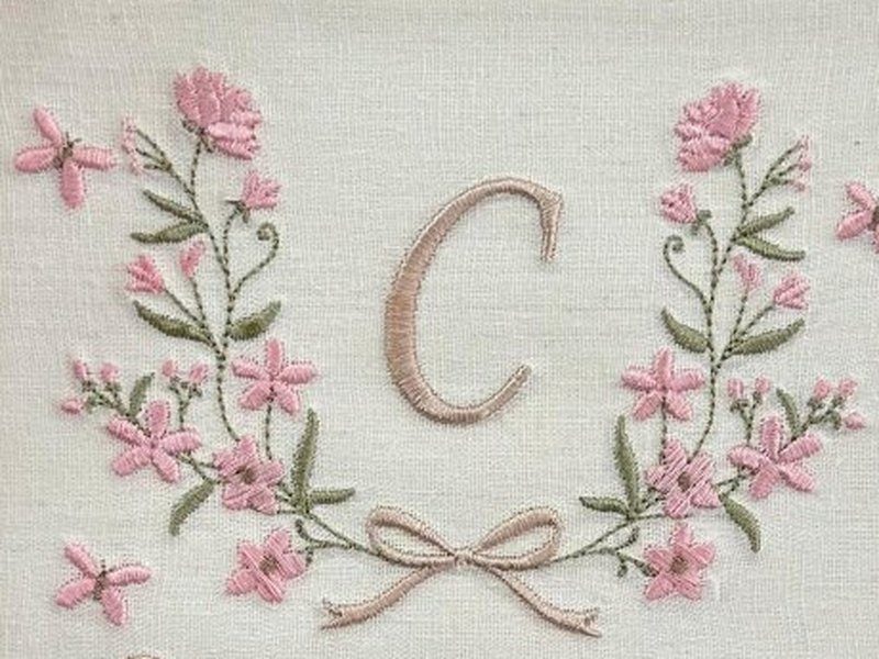 Coroa de flores rosa e folhas verdes em forma de C aberto, com a letra C bege ao centro e um la�o bege na base