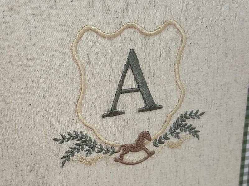 Escudo de contorno creme com a letra A verde escuro, dois ramos de louro e um cavalinho de balan�o marrom bordado na base