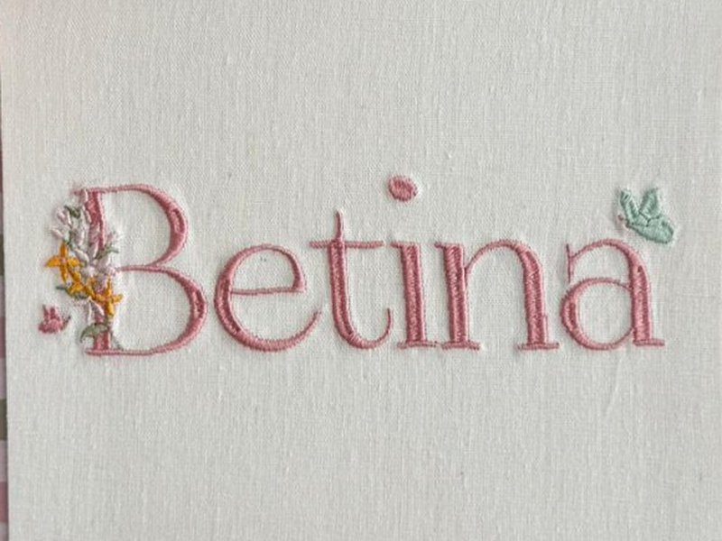 O nome Betina bordado por inteiro em letra serifada rosa, com um buqu� mi�do de flores brancas e amarelas saindo da letra B