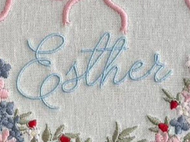 O nome Esther bordado por inteiro em cursiva azul-clara, no centro da coroa