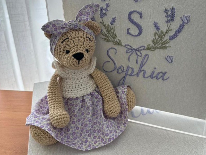 Ursa de croch� bege com vestido de tecido floral lil�s, gola branca de croch� e len�o lil�s amarrado na cabe�a