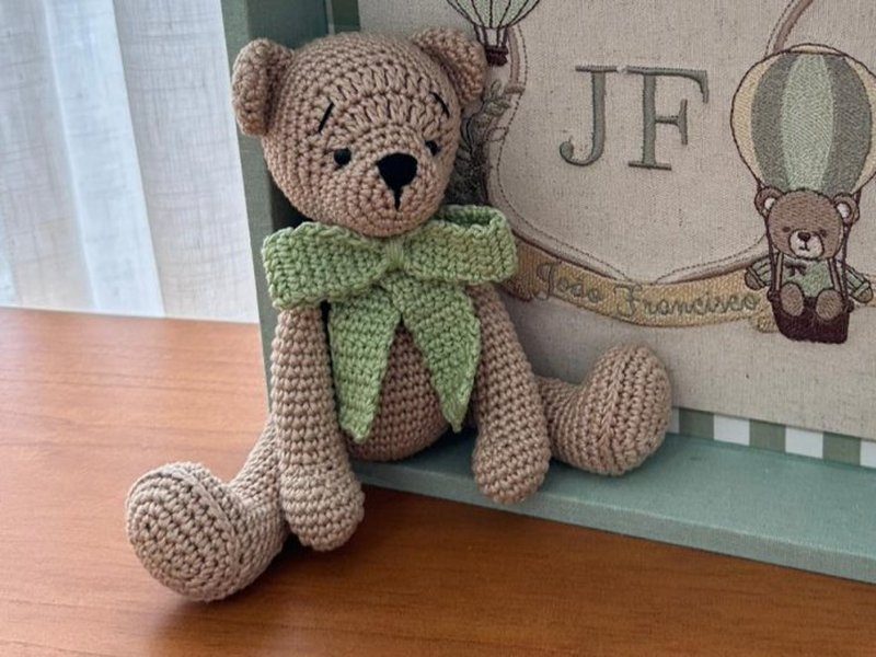 Urso de croch� bege com la�o de croch� verde no pesco�o, sentado dentro da caixa com as pernas para fora da moldura