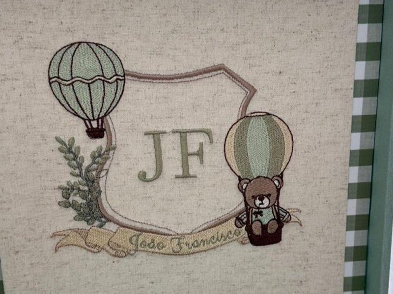 Escudo bege com 'JF' verde escuro, um bal�o listrado � esquerda e outro � direita levando um ursinho de gravata na cesta, ramo de folhas e faixa com 'Jo�o Francisco'