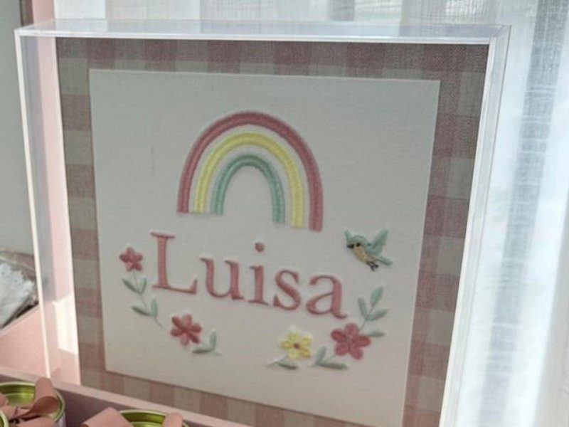 O quadro acr�lico com o arco-�ris bordado em rosa, amarelo e verde, 'Lu�sa' em rosa, flores e um beija-flor