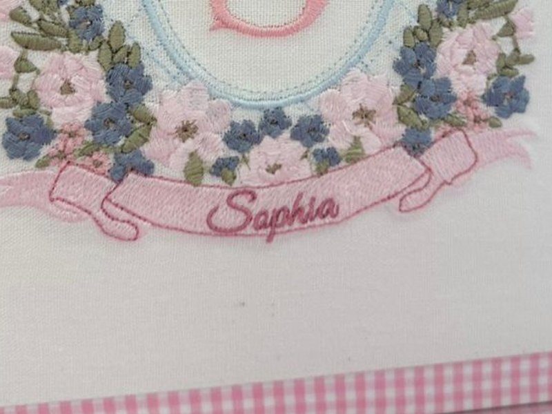 'Sophia' bordado em cursiva sobre a faixa rosa, com as pontas dobradas da fita
