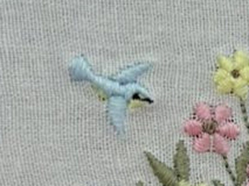 Um passarinho azul bordado em relevo, em voo, � esquerda da coroa de flores