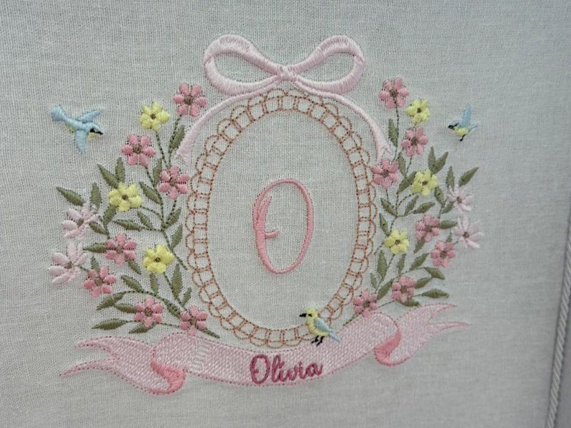 Oval formado por uma correntinha de p�rolas bege com a letra O rosa ao centro, la�o rosa no topo e coroa de flores rosas e amarelas